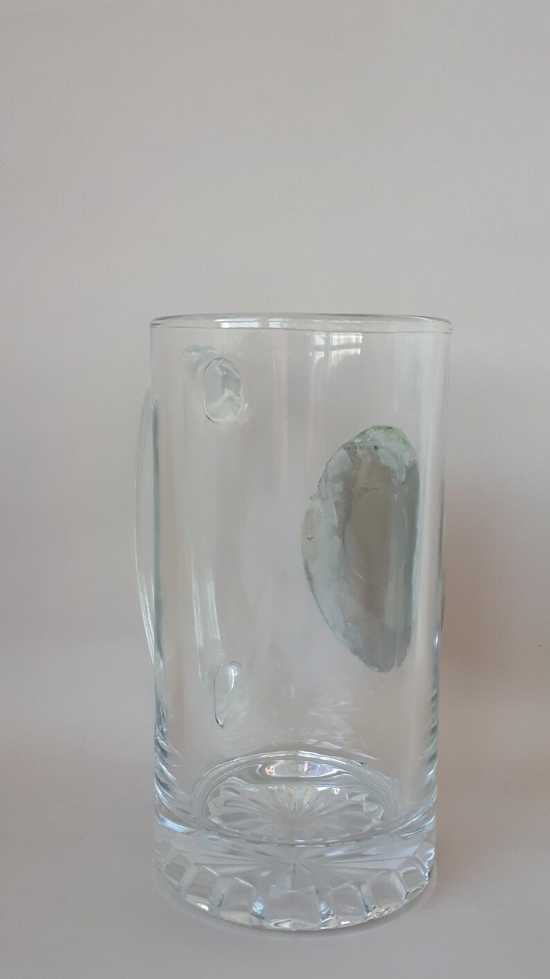 May include: A clear glass beer mug with a handle. The mug has a decorative, cut-glass base and a cylindrical body. There are a few small imperfections on the glass. The mug is empty and sits against a plain background.