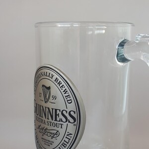 May include: Clear glass Guinness beer mug with a handle. The mug features the Guinness logo on the side, including the harp symbol and the words "Extra Stout" and "Dublin".