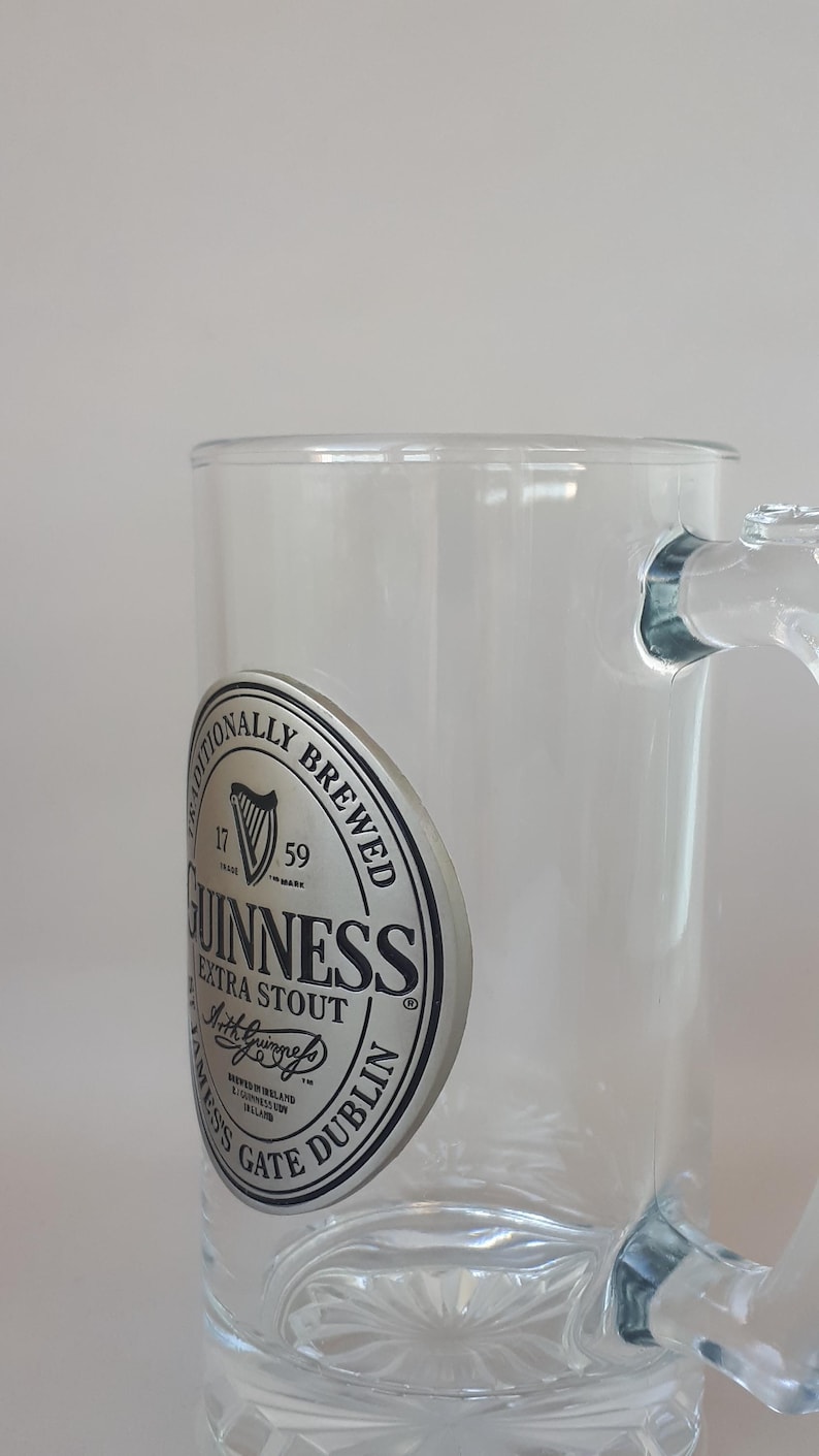 May include: Clear glass Guinness beer mug with a handle. The mug features the Guinness logo on the side, including the harp symbol and the words "Extra Stout" and "Dublin".