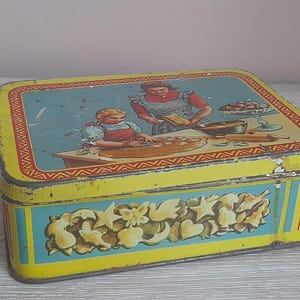 Op de afbeelding: Vintage gele blikken doos met een blauw deksel met een bakscène. De doos heeft een decoratief bloemmotief aan de voorkant en een rood-gele rand. Een charmante opbergdoos.