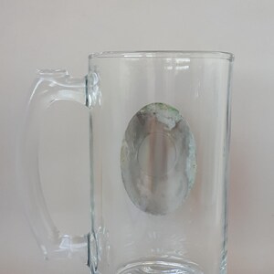 May include: A clear glass beer mug with a handle. The mug has a decorative base and a faded, oval-shaped design on the front. The handle is clear and attached to the side of the mug. The mug is empty.