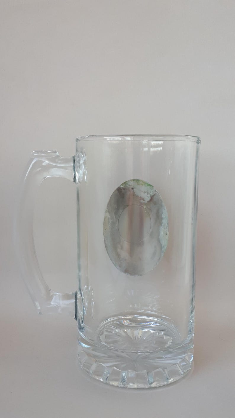 May include: A clear glass beer mug with a handle. The mug has a decorative base and a faded, oval-shaped design on the front. The handle is clear and attached to the side of the mug. The mug is empty.