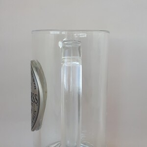 May include: Clear glass beer mug with a handle. The mug has a decorative base and a silver and black emblem on the side. The emblem has the word "Guinness" on it.