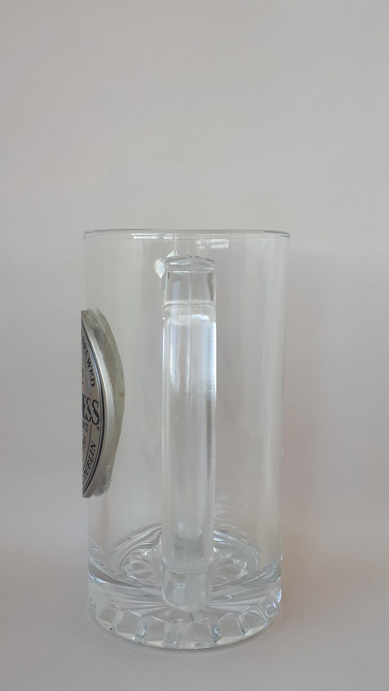 May include: Clear glass beer mug with a handle. The mug has a decorative base and a silver and black emblem on the side. The emblem has the word "Guinness" on it.