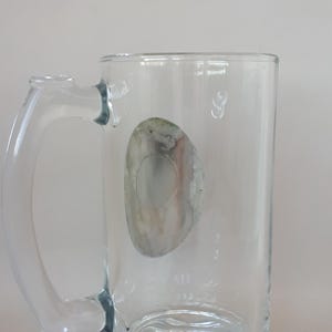 May include: Clear glass beer mug with a handle. The mug has a decorative base and a faded, oval-shaped sticker on the side. The handle is curved and attached to the side of the mug. The mug is empty.