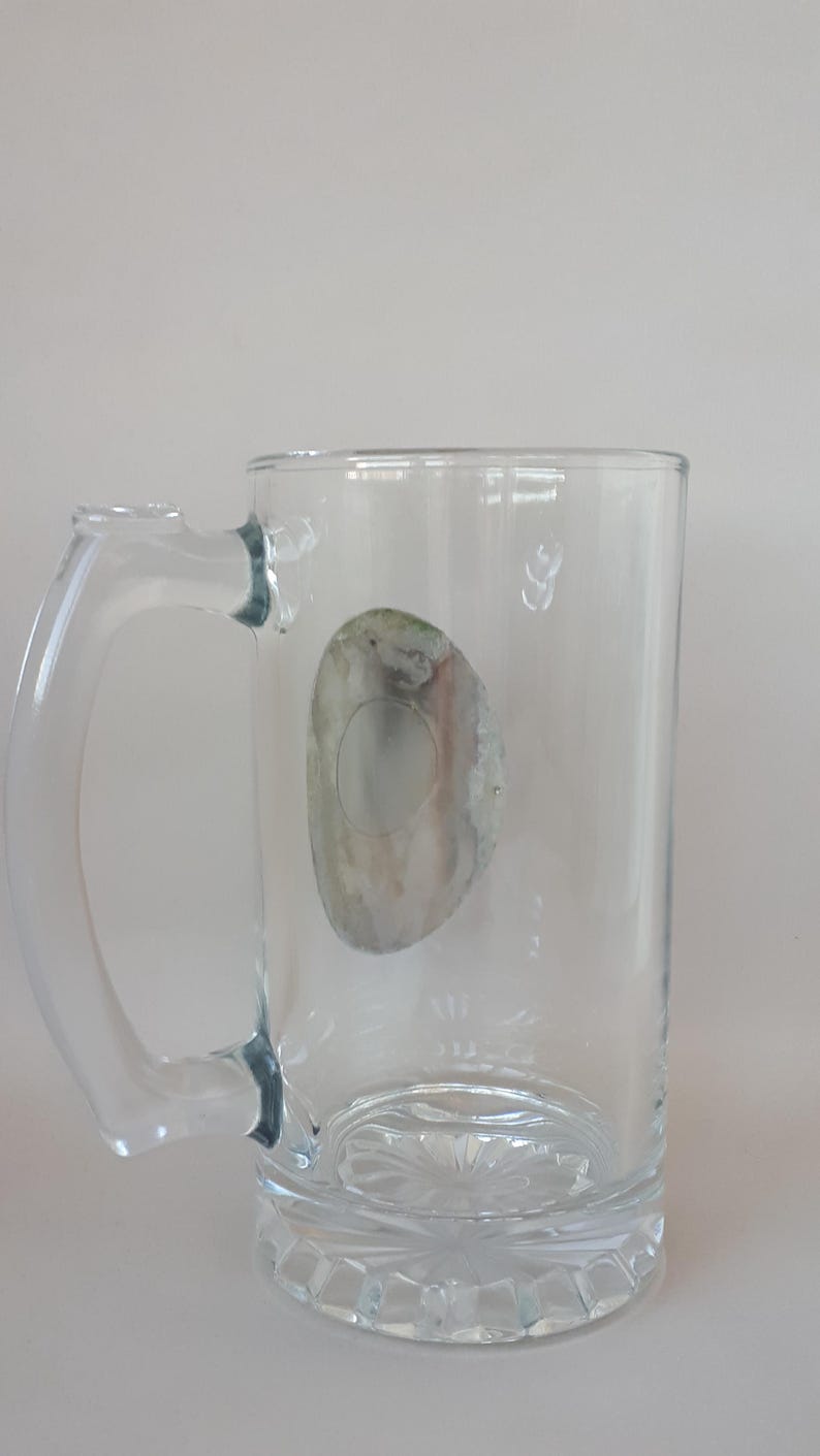 May include: Clear glass beer mug with a handle. The mug has a decorative base and a faded, oval-shaped sticker on the side. The handle is curved and attached to the side of the mug. The mug is empty.