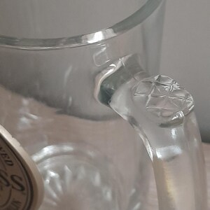 May include: Clear glass beer mug with a handle. The handle has a diamond pattern on the top. The mug is empty and appears to be clean. The glass is thick and sturdy, suitable for holding cold beverages.
