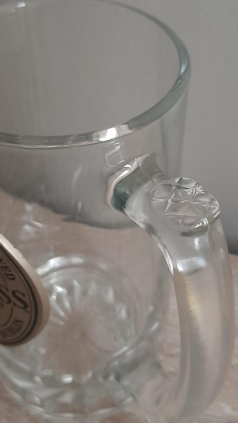 May include: Clear glass beer mug with a handle. The handle has a diamond pattern on the top. The mug is empty and appears to be clean. The glass is thick and sturdy, suitable for holding cold beverages.