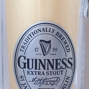 May include: A clear glass beer mug with a handle, filled with a light-colored liquid. The mug features a silver and black Guinness Extra Stout logo with the text "Traditionally Brewed" and "St James's Gate Dublin."