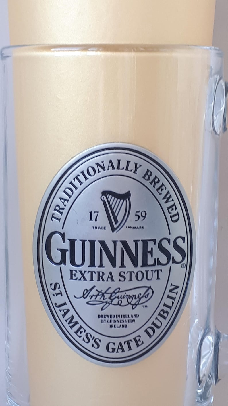 May include: A clear glass beer mug with a handle, filled with a light-colored liquid. The mug features a silver and black Guinness Extra Stout logo with the text "Traditionally Brewed" and "St James's Gate Dublin."