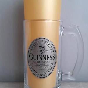 May include: A clear glass beer mug with a handle, featuring a gold-colored interior and a gold-colored top. The mug has a silver label with the Guinness Extra Stout logo and text. The mug is a classic beer drinking vessel.