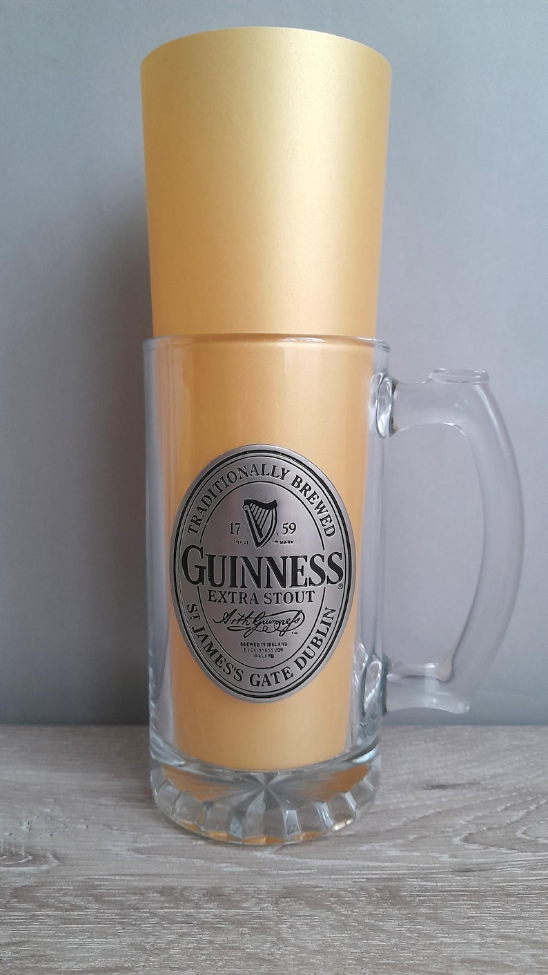May include: A clear glass beer mug with a handle, featuring a gold-colored interior and a gold-colored top. The mug has a silver label with the Guinness Extra Stout logo and text. The mug is a classic beer drinking vessel.