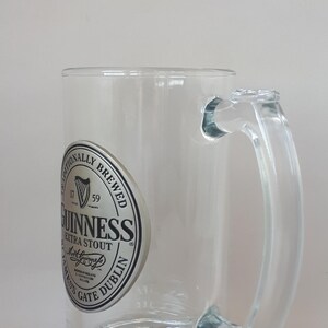 May include: Clear glass beer mug with a handle. The mug features the Guinness Extra Stout logo on the front. The base of the mug has a decorative pattern.