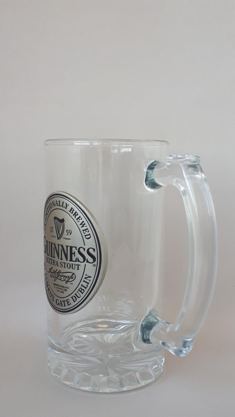 May include: Clear glass beer mug with a handle. The mug features the Guinness Extra Stout logo on the front. The base of the mug has a decorative pattern.