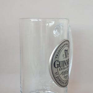 May include: A clear, cylindrical Guinness beer glass with a handle. The glass features a silver emblem with the Guinness logo and the words "Extra Stout" and "St. James's Gate". The base of the glass has a decorative, starburst design.