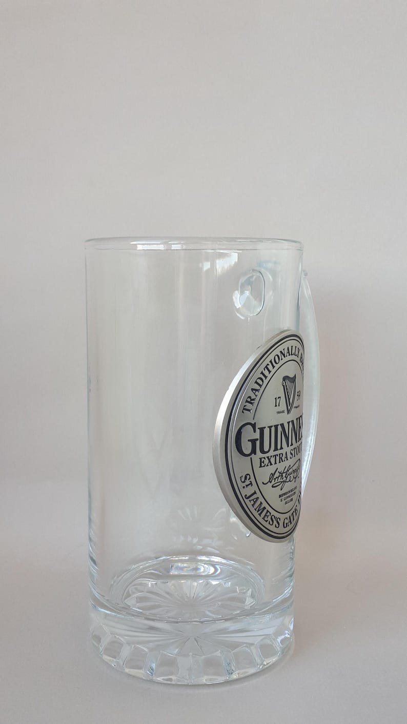 May include: A clear, cylindrical Guinness beer glass with a handle. The glass features a silver emblem with the Guinness logo and the words "Extra Stout" and "St. James's Gate". The base of the glass has a decorative, starburst design.