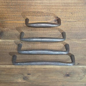 May include: Four hand-forged, dark gray metal cabinet pulls of varying lengths are arranged vertically on a wooden surface. Each pull has a curved handle with a slight texture, and the ends are bent upwards.