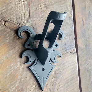 May include: Black metal wall mount bracket with a decorative design. The bracket has a curved shape and a hole for mounting.