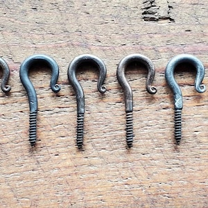 May include: Seven antique-style metal screw-in hooks with a dark, aged finish. The hooks are arranged in a row on a weathered wooden surface, showcasing their curved design and threaded base for easy installation.