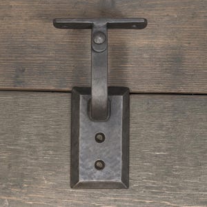 May include: A dark gray metal bracket with a rectangular base and a horizontal bar extending from the top. Two circular holes are visible on the base. The bracket is mounted on a wooden surface.
