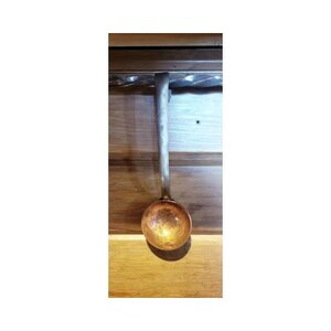 May include: A copper-colored ladle with a long, silver-toned handle. The round bowl of the ladle is a warm, metallic orange. The handle is attached to a wooden surface.