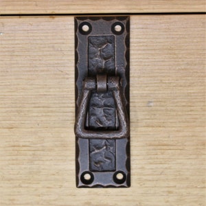 May include: A dark bronze metal drawer pull with a textured surface and a rectangular backplate. The pull has a hinged handle and is mounted on a light wood surface. The hardware features four screw holes.