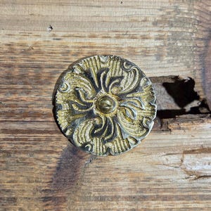 May include: Ornate, circular metal furniture hardware with a gold and silver finish. The decorative piece features a central button and a detailed floral design. The hardware is set against a rustic wooden surface.
