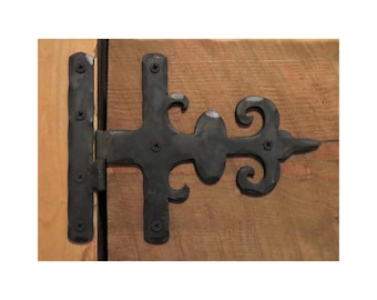 May include: Black wrought iron hinge with decorative scrollwork and a fleur-de-lis design. The hinge is attached to a wooden surface with screws. The hinge is a decorative element for doors or gates.