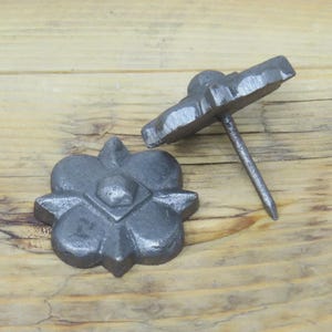 May include: Two cast iron decorative nail heads. Each nail head is shaped like a four-petal flower with a square center. One nail head is lying flat, while the other has a nail extending from the back. The nails are a dark gray color.