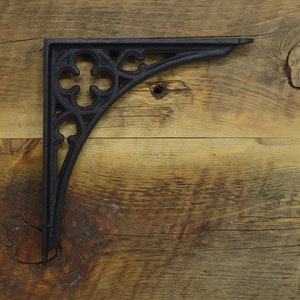 Op de afbeelding: Zwarte gietijzeren plankbeugel met een sierlijk, gotisch design. De beugel heeft een decoratief klaverbladpatroon en een haakse vorm, geschikt voor het ondersteunen van planken. De beugel wordt getoond tegen een rustieke houten achtergrond.