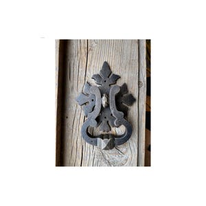 May include: A black metal door knocker with a decorative, ornate design. The knocker is attached to a wooden door.