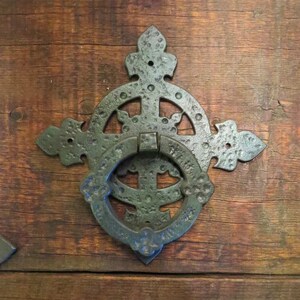 May include: A black metal door knocker with a circular design and a ring handle. The knocker is attached to a wooden door.