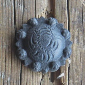 May include: A dark gray, decorative metal rosette with an ornate, swirling design. The rosette is affixed to a weathered, vertical wooden surface. The rosette has a scalloped edge with small, round embellishments.