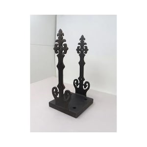 May include: A black metal decorative stand with two ornate, vertical supports and a square base. The supports feature a fleur-de-lis design at the top and decorative scrollwork. The stand is designed to hold an item.