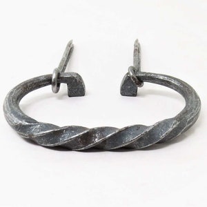 Op de afbeelding: Een gedraaide metalen ring met twee metalen pinnen die uit de bovenkant steken. De ring is donker zilverkleurig en heeft een ruwe, gehammerde textuur.