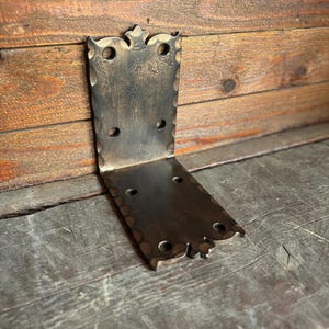 May include: A dark, L-shaped metal bracket with decorative edges and multiple holes. The bracket has a rustic, aged appearance, suggesting it is vintage or handcrafted. It is designed to be mounted on a corner.