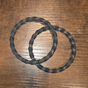 May include: Two circular bracelets crafted from dark gray metal, each featuring a continuous twisted rope-like texture. The bracelets are linked together, showcasing a uniform matte finish across the entire surface of the metal bands.