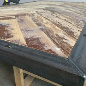 May include: A rectangular table featuring a light brown wooden top arranged in a herringbone pattern. The surface is enclosed by a dark gray metal frame with visible welded corner joints and metal rivets secured along the perimeter edges.