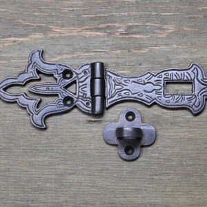 May include: Black metal latch with ornate details and a decorative hinge. The latch has a rectangular opening and a small, separate hook.