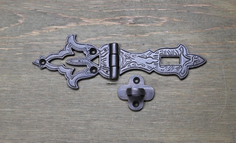 May include: Black metal latch with ornate details and a decorative hinge. The latch has a rectangular opening and a small, separate hook.