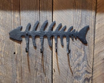 May include: Black metal fish skeleton wall hook with five hooks. The hook is mounted on a rustic wooden wall.