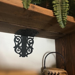 May include: Black metal shelf bracket with a decorative scroll design. The bracket is attached to a wooden shelf and a white wall. A small basket with a fabric liner featuring a black bunny rabbit design is on the shelf below the bracket.