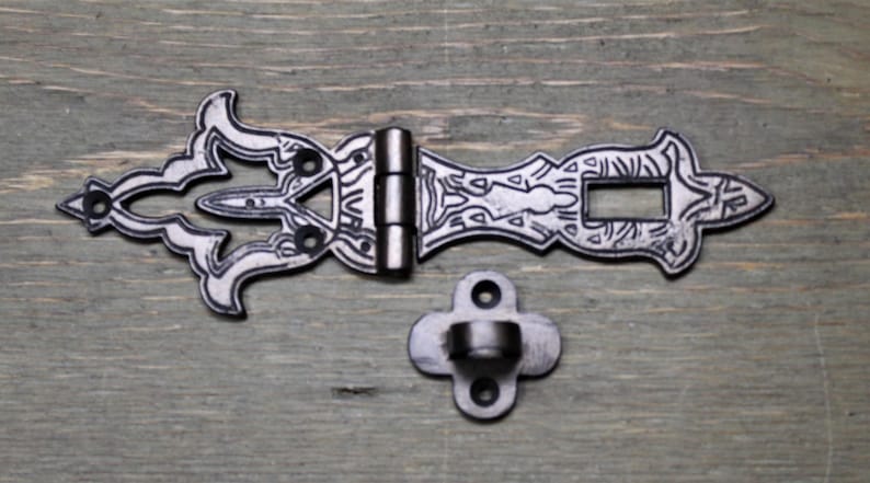 May include: A silver-toned metal latch with an ornate design. The latch has a hinge and a separate mounting plate. The latch is designed to be used on a door or cabinet.