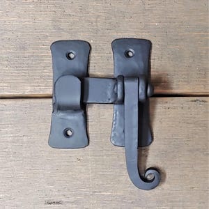 May include: Black metal door latch with a decorative scroll design. The latch is mounted on a wooden surface.