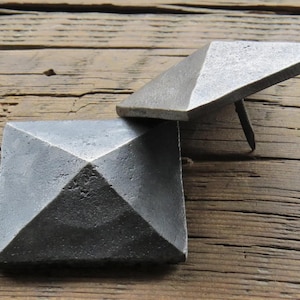 May include: Two square pyramid-shaped metal nails with a silver finish. The nails are lying on a wooden surface.