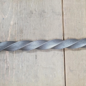 May include: A twisted metal rod with a textured, gray finish. The rod is made of a series of overlapping, curved sections, creating a spiral design. The rod is set against a light brown wooden surface.