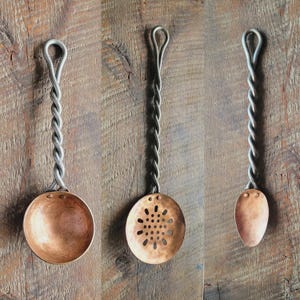 May include: Three handcrafted kitchen utensils with copper bowls and twisted metal handles. The set includes a ladle, a slotted spoon, and a small spoon. The handles are silver-colored, and the bowls have a warm copper hue. The utensils are displayed against a wooden background.