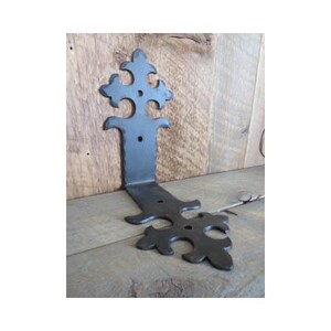 May include: A black, decorative metal bracket with an ornate, fleur-de-lis design. The L-shaped bracket is made of wrought iron and features a textured surface. The bracket is approximately 6 inches tall and 4 inches wide.