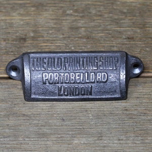 Puede incluir: Un tirador de cajón de metal negro con el texto "THE OLD PRINTING SHOP PORTOBELLO RD LONDON" grabado en él.