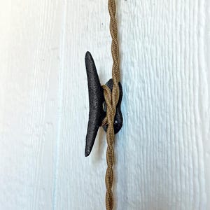 May include: A black cleat-shaped hook with a brown twisted cord, mounted on a white wooden surface. The hook is made of cast iron and is designed to hold the cord in place. The cord is a decorative element.